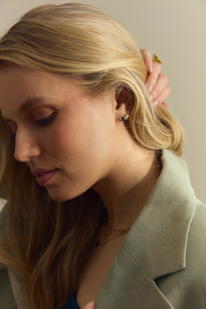 A close-up image of a woman's ear wearing a gold stud earring with a blue and green cushion-cut sapphire.