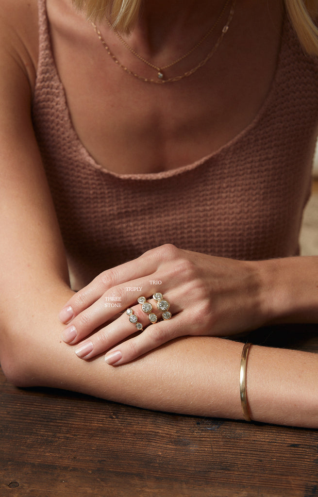 close up of woman wearing gold ring with three bezel set round white diamonds alongside other diamond rings
