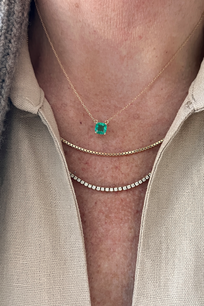 close up of woman wearing cushion cut colombian emerald necklace alongside other gold necklaces