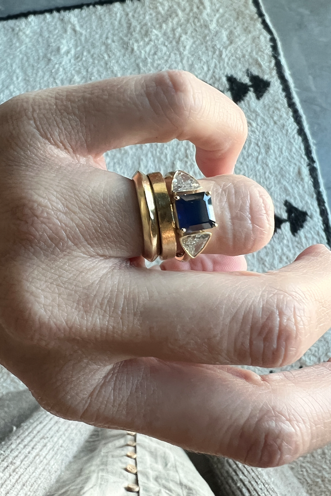 close up of hand wearing sapphire gold ring with trillion white diamonds alongside other gold bands