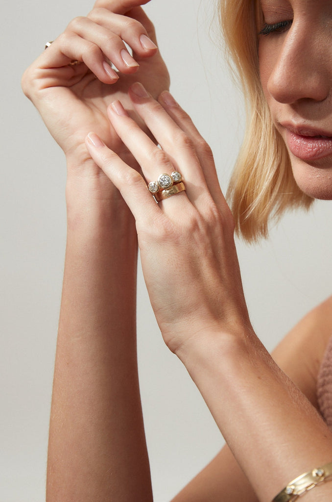 woman wearing gold ring with three bezel set round white diamonds alongside other gold jewelry