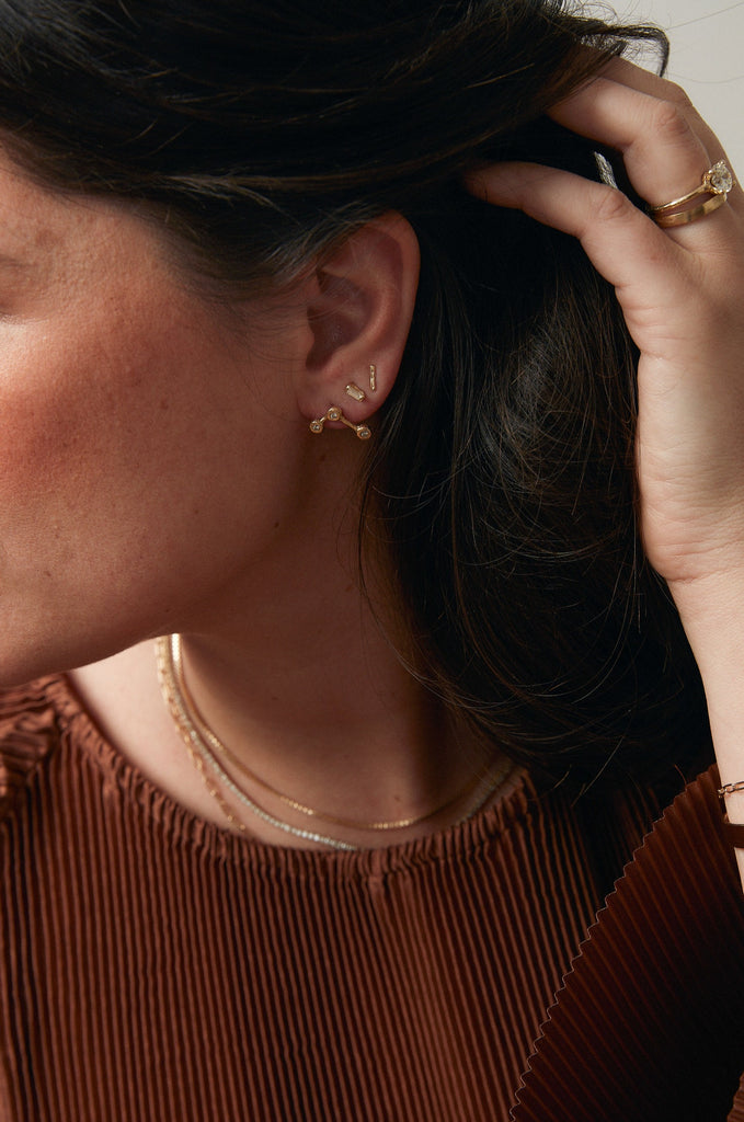 woman wearing gold constellation inspired stud earring with three round white diamonds alongside other gold jewelry