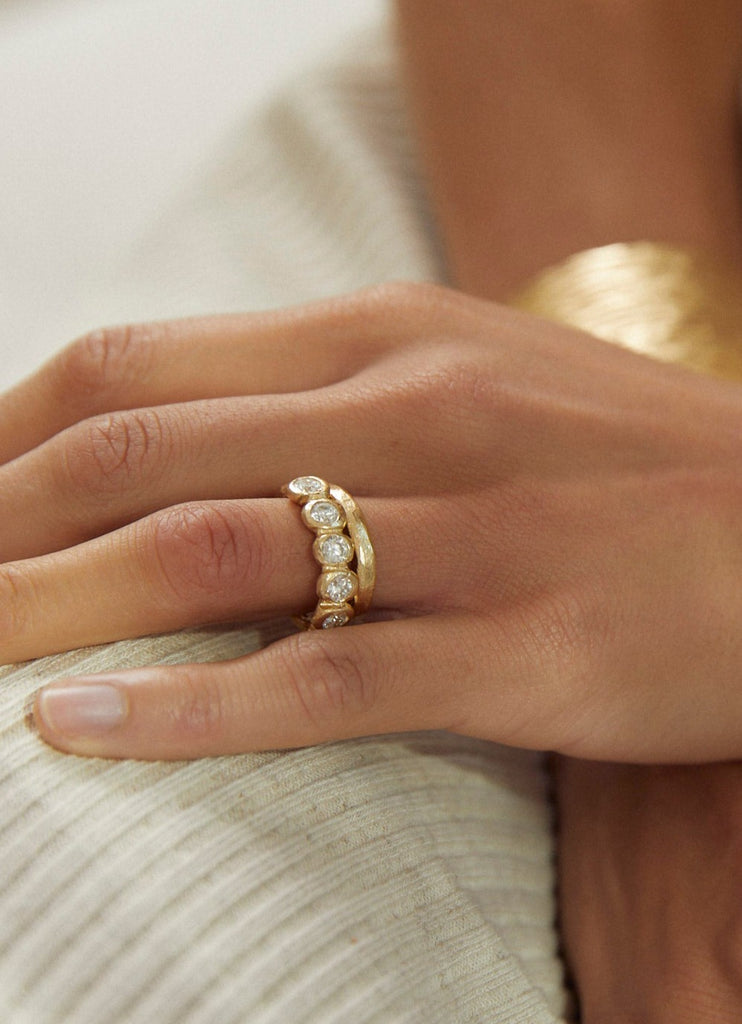 close up of woman wearing gold band with six bezel set round diamonds alongside other gold jewelry