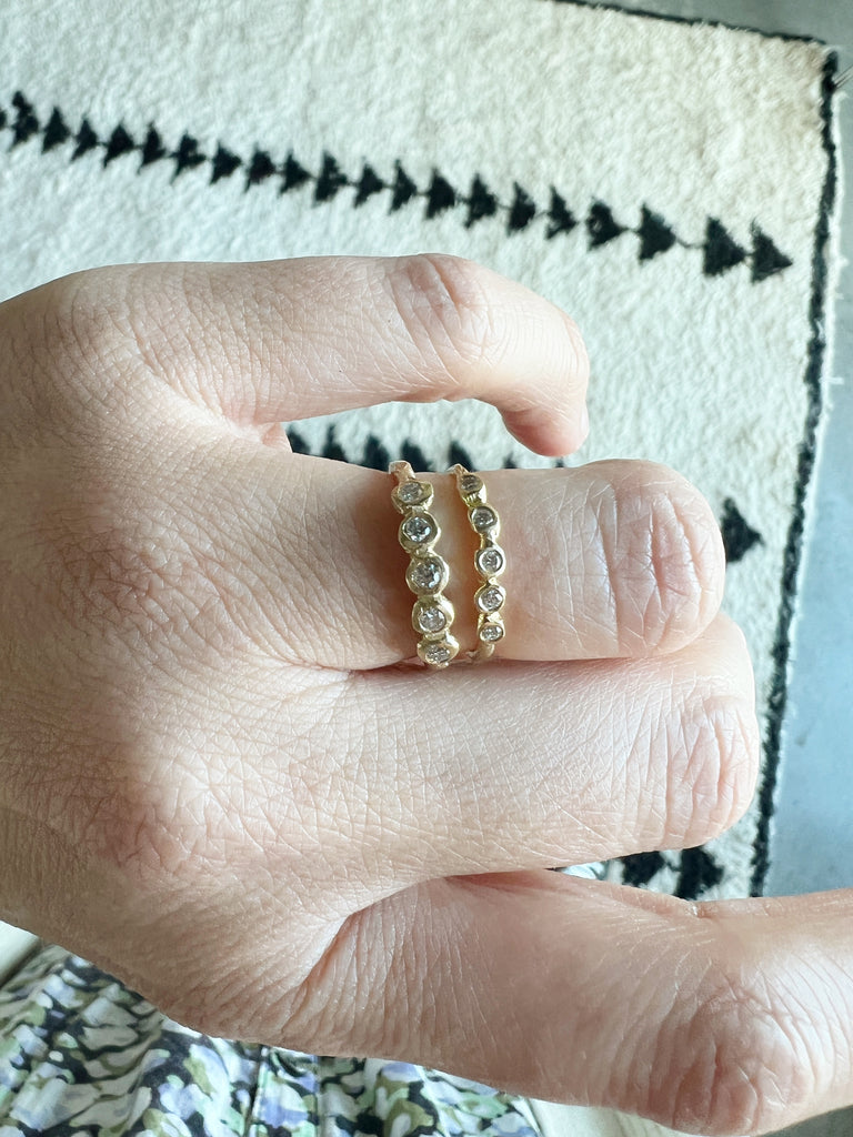 close up of hand wearing gold ring with five bezel set round white diamonds alongside other gold band