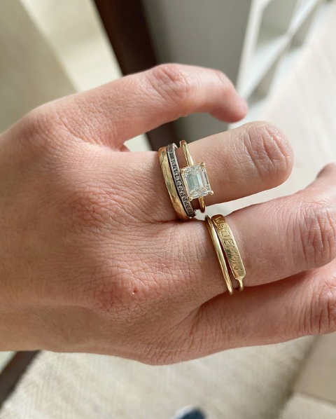 close up of hand wearing gold four prong emerald cut white diamond ring alongside other rings