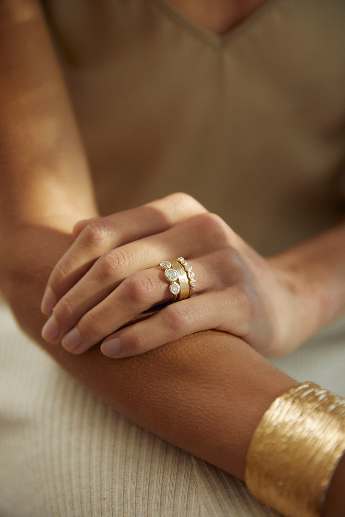 close up of woman wearing gold ring with five bezel set round white diamonds alongside other gold jewelry