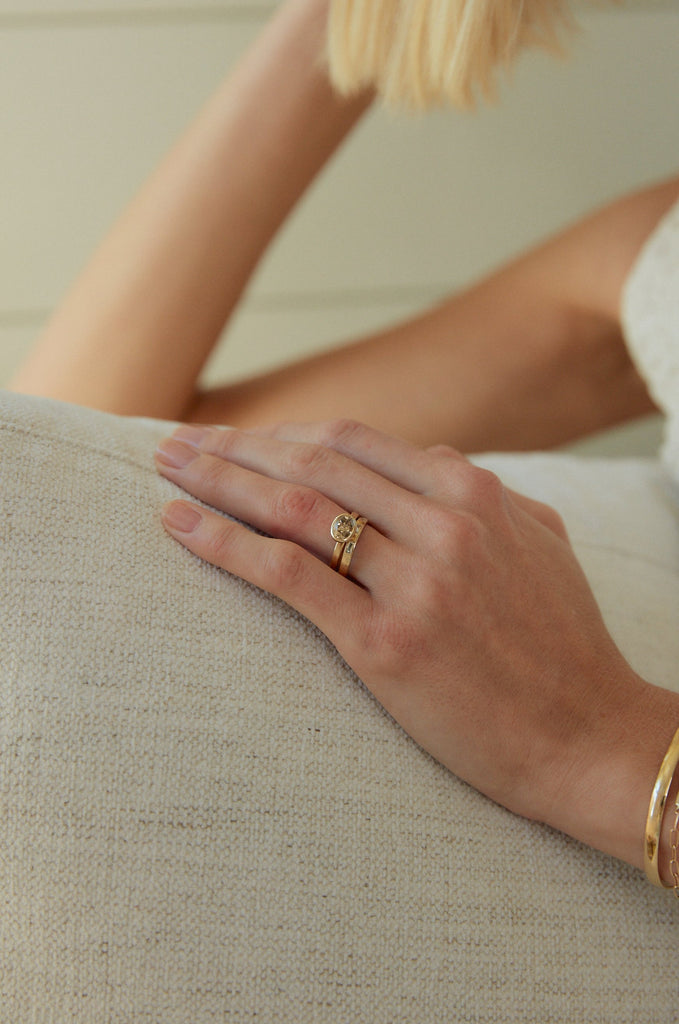 close up of woman's hand wearing gold ring with bezel set oval champagne diamond alongside other gold jewelry