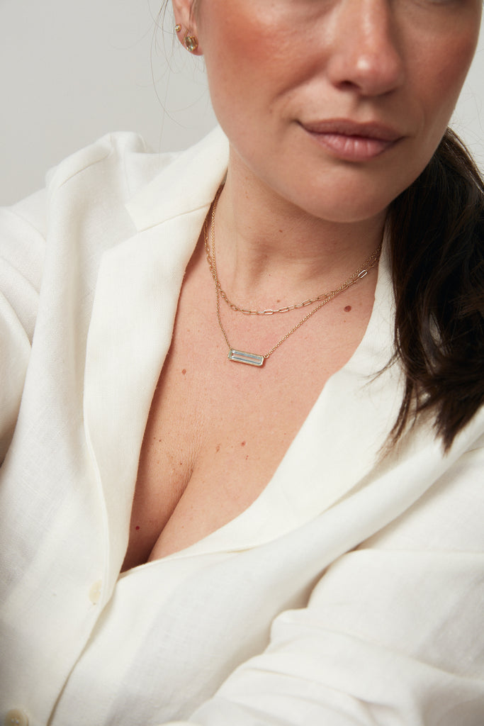 close up of woman wearing gold necklace with elongated emerald cut aquamarine bar alongside other gold jewelry