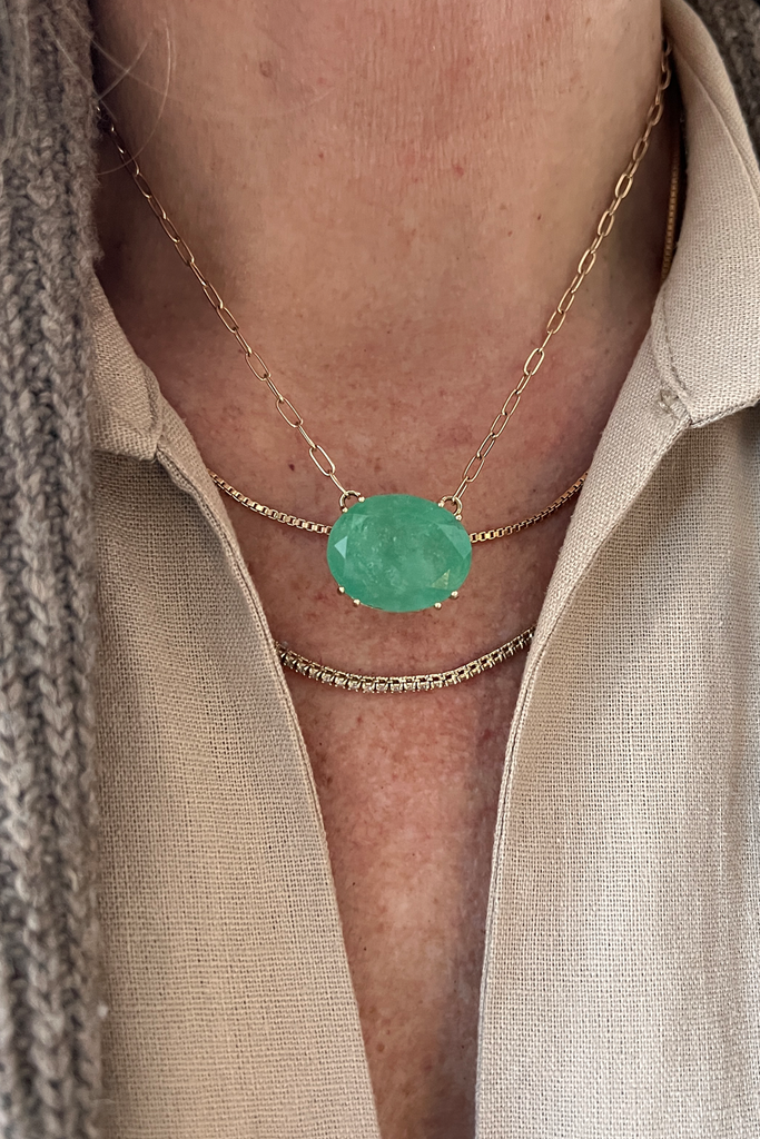 close up of woman wearing oval colombian emerald necklace on gold cable chain alongside other gold necklaces