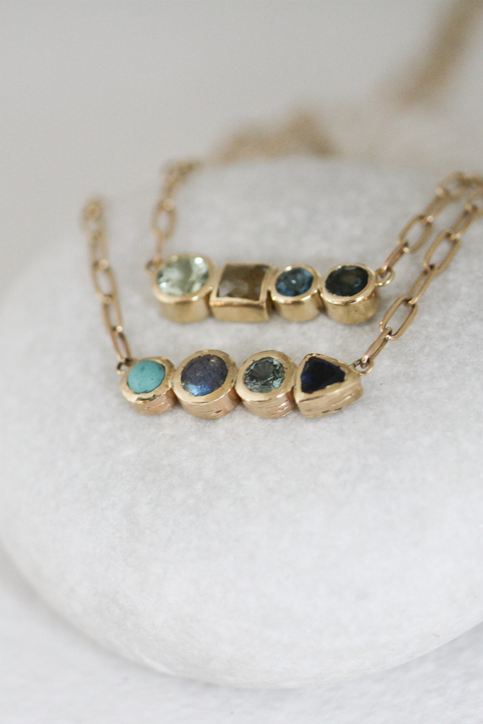 close up of gold necklace with round green amethyst, princess rosecut mustard diamond, round cerulean topaz, and round bluegreen sapphire alongside another four stone necklace