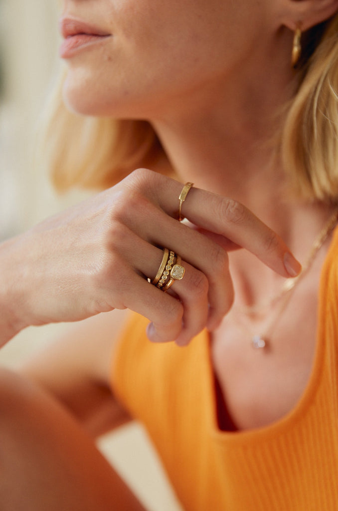 close up of woman wearing gold ring with cushion cut bezel set white diamond alongside other gold rings