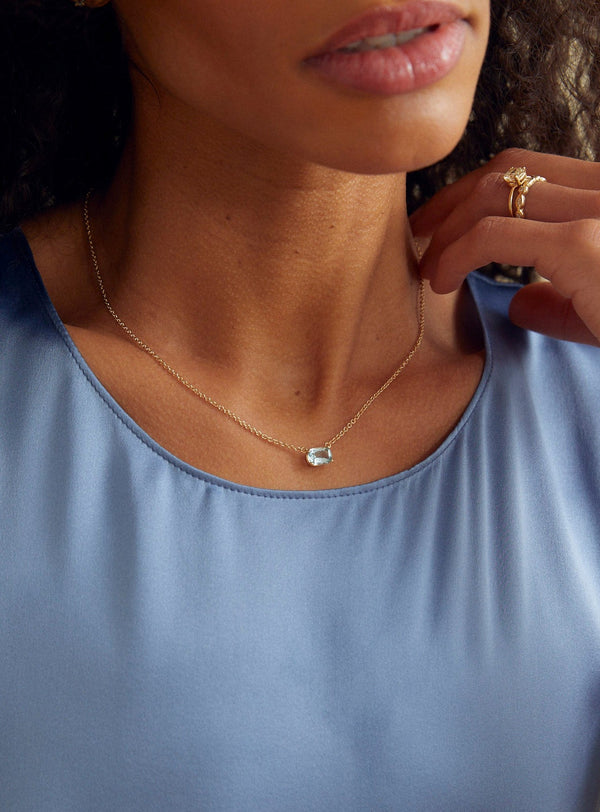 close up of woman wearing gold necklace with cushion cut rectangle aquamarine alongside other gold jewelry 