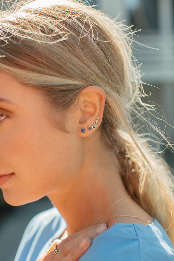 close up of woman wearing gold stud with trillion sapphire alongside other gold earrings