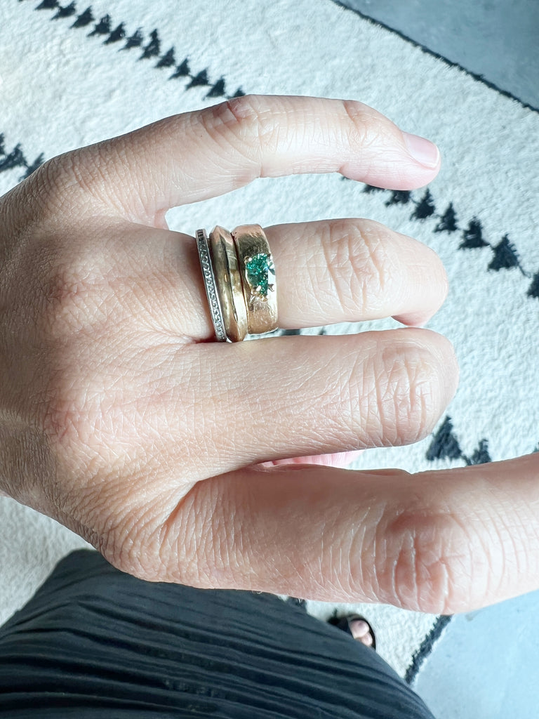 close up of hand wearing wide gold band with blue green pear diamond alongside other rings