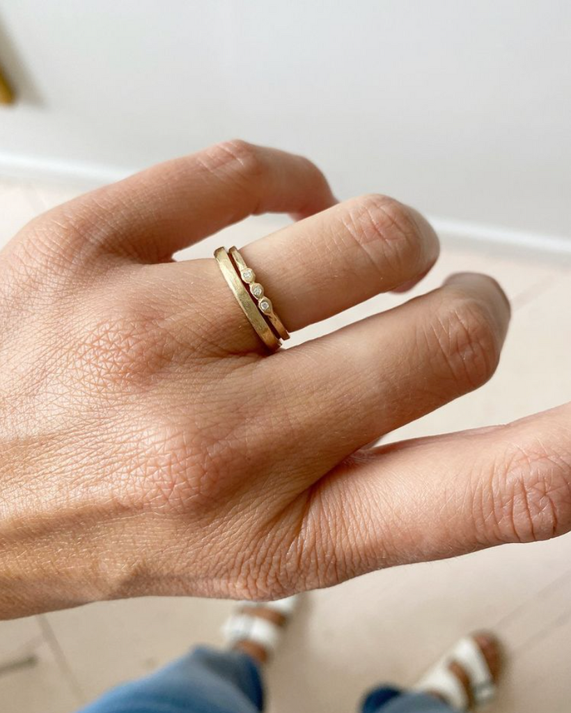 close up of hand wearing thin gold band with three bezel set round white diamonds alongside other gold band