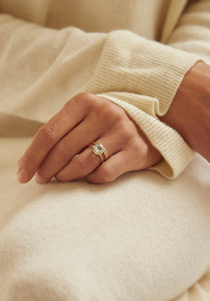close up of woman wearing gold four prong ring with textured band and cushion cut aquamarine alongside other gold rings