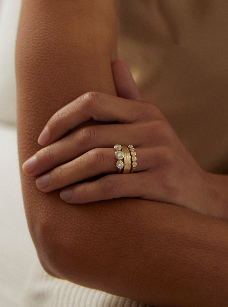 close up of woman wearing gold ring with three bezel set round white diamonds alongside other gold rings